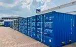 Reihe blauer Frachtcontainer auf gepflasterter Fläche bei Lager-Ein DE GmbH Schwülper unter bewölktem Himmel. Gebäudedach im Hintergrund.