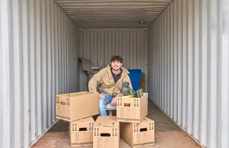 Mann sitzt in einem leeren Lagercontainer von Sirius mit Umzugskartons, beschriftet mit Umzugskarton.