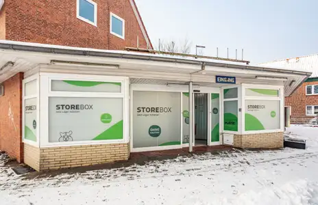 Außenansicht von Storebox in Stade mit beschrifteten Schaufenstern und Eingang Schild, umgeben von verschneiter Landschaft.