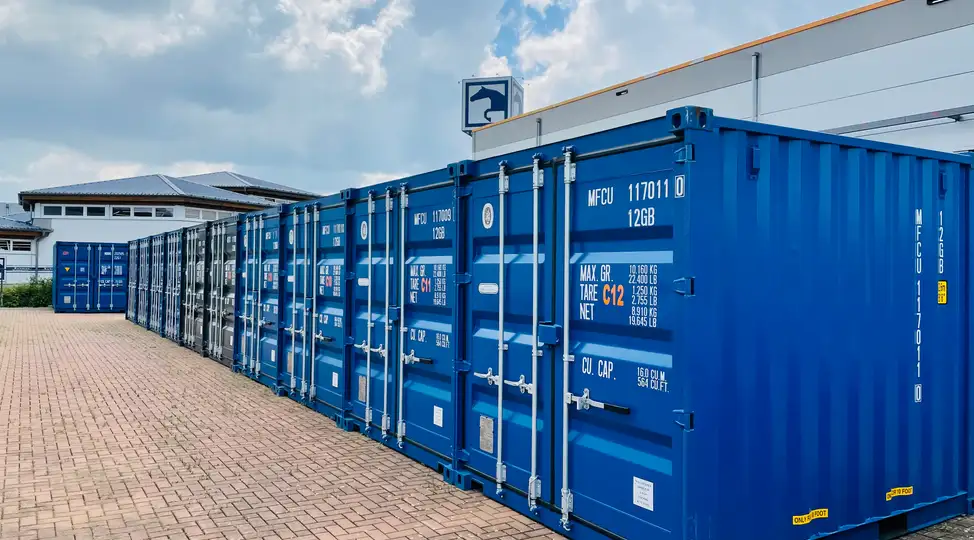 Reihe blauer Frachtcontainer auf gepflasterter Fläche bei Lager-Ein DE GmbH Schwülper unter bewölktem Himmel. Gebäudedach im Hintergrund.