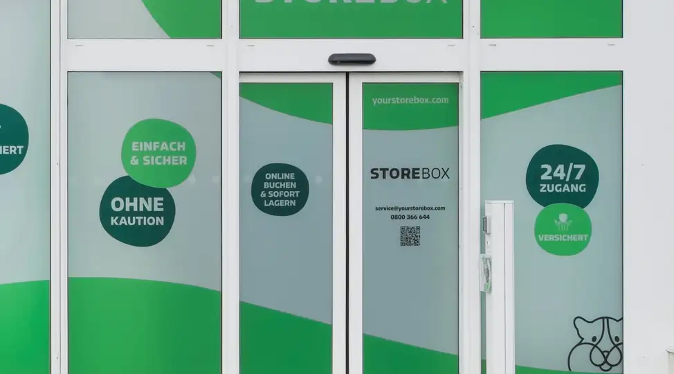 Außenansicht eines Storebox-Standorts mit grüner Fensterfolie, beschriftet mit Einfach & Sicher, Ohne Kaution und 24/7 Zugang.