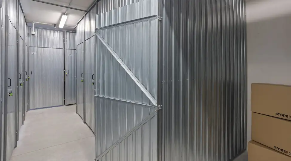 Lagerraum bei Storebox Recklinghausen: Metallene Lagerabteile, geöffnete Tür im Vordergrund, Kartons am Boden.